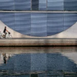 Dwóch rowerzystów jedzie elektrycznymi rowerami wzdłuż nowoczesnego budynku, odbicie w wodzie